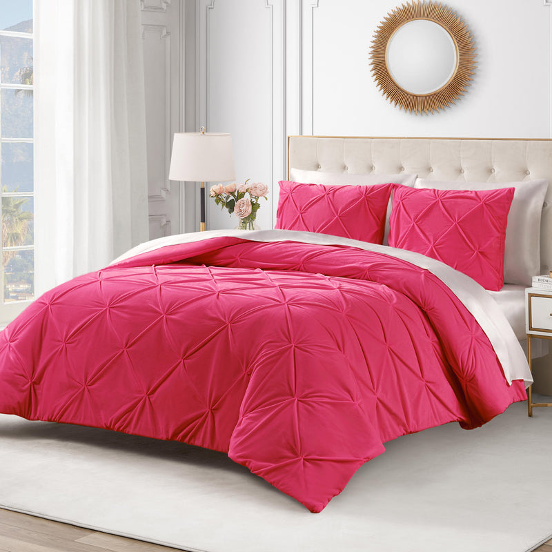 A neatly made bed features the Kiss Pleat Micromink Comforter Set—luxurious, textured, and bright pink—paired with matching pillows in a white bedroom with a tufted beige headboard, round mirror, lamp, and flowers on the nightstand.