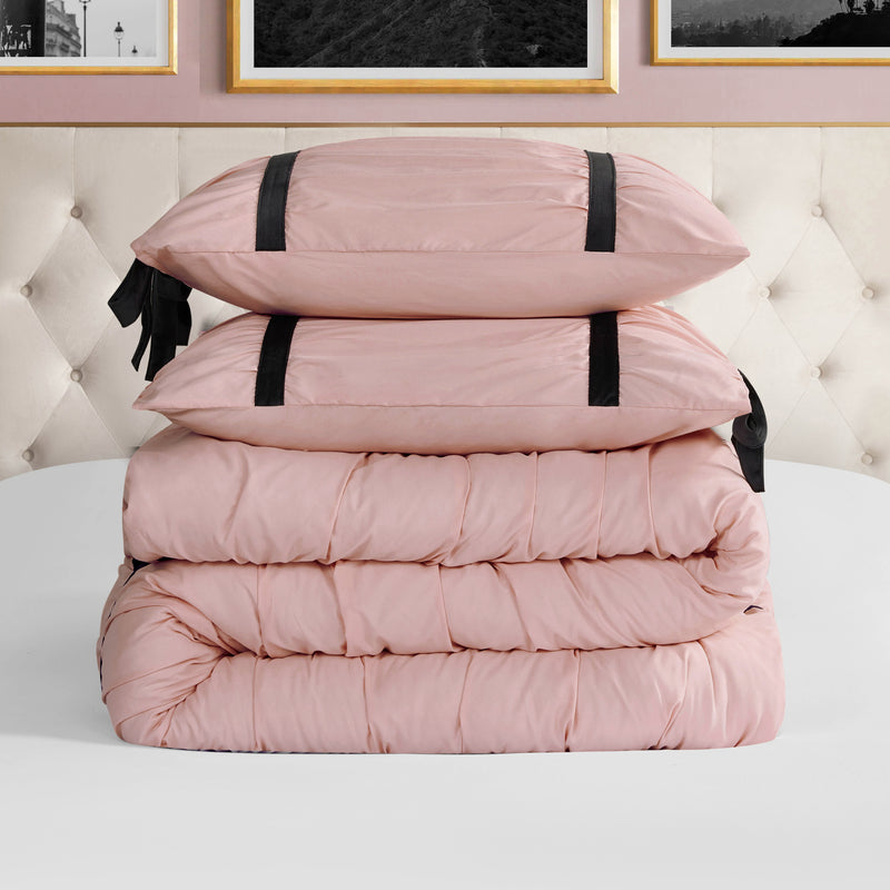 A neatly folded light pink Amelie Bows Ruched Comforter Set with two matching pillows featuring black velvet bows sits on a white surface before a tufted beige headboard and framed wall art.