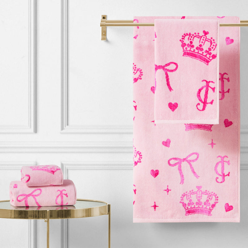 The Printed Jacquard Bath Towel Set, featuring bright pink crown, heart, bow, and letter designs, hangs on a gold rack with matching towels folded on a round gold table against a white paneled wall.