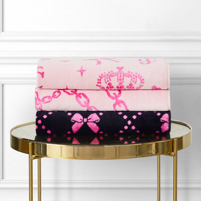 Two Printed Jacquard Bath Towels—one light pink with crowns and chains, one black with pink bows and diamonds—are stacked on a round gold side table against a white paneled wall.