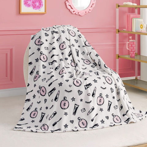 A Kids Plush Throw Blanket featuring pink and black makeup-themed illustrations—perfume bottles, lipsticks, eyelashes, and stars—is draped over a chair in a pink room with wall art and shelves in the background.