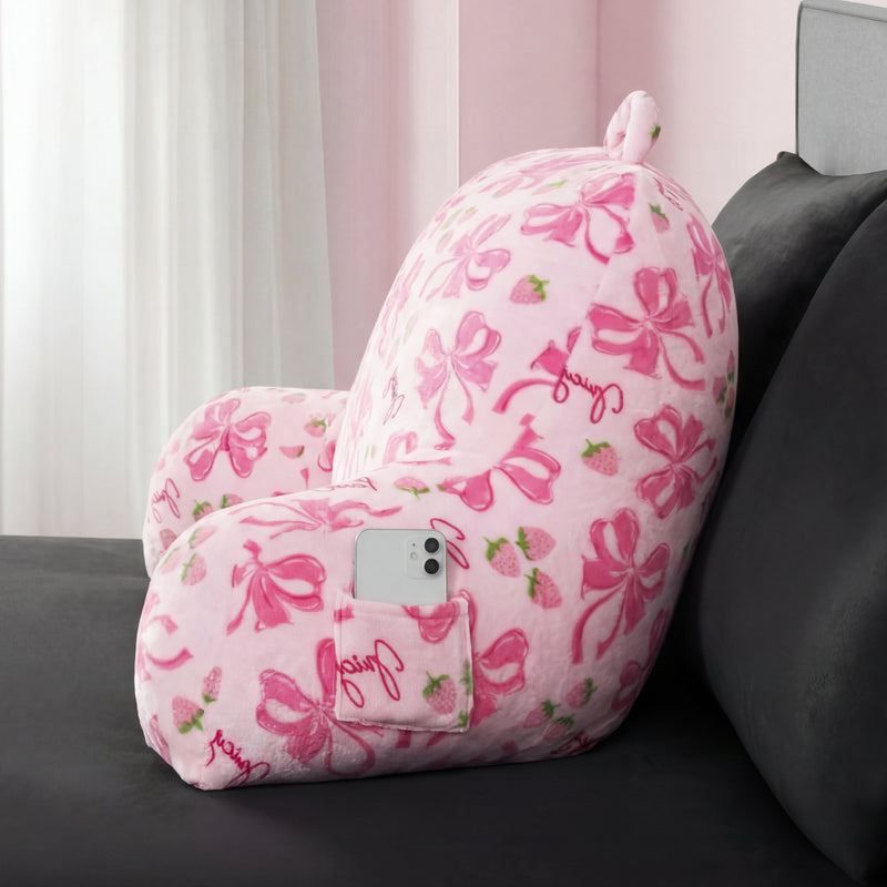 The Backrest Pillow, a plush pink pillow with phone pocket, armrests, pink bows, and strawberries, sits on a dark gray bed. A smartphone rests in its side pocket while sunlight streams through the nearby window.