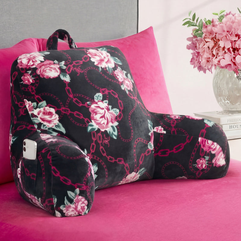 A Backrest Pillow with armrests, featuring black fabric with pink floral and chain designs, sits on a pink bed. A smartphone is stored in its side pocket, and a vase of pink flowers is placed on the nearby nightstand.
