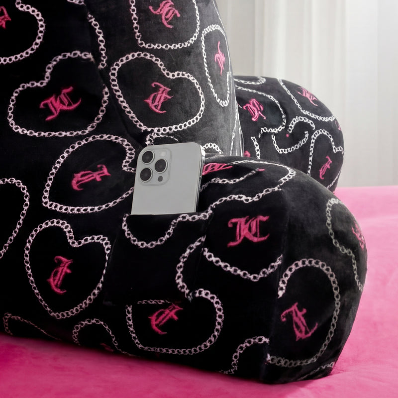 The Backrest Pillow features pink heart and chain patterns, a small pocket holding a silver smartphone, and sits on a pink bedspread with a white curtain in the background.