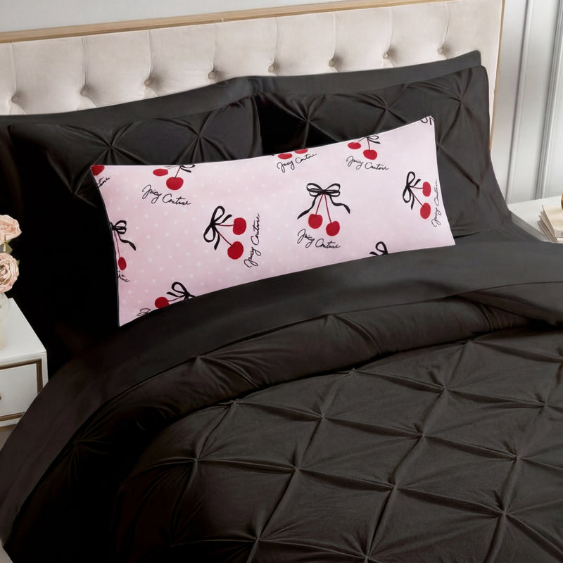 A bed with a black tufted comforter and matching pillowcases features the Plush Body Pillow—oversized, plush, and pink with red cherries, black bows, and cursive accents. The chic pattern pops against the beige tufted headboard.