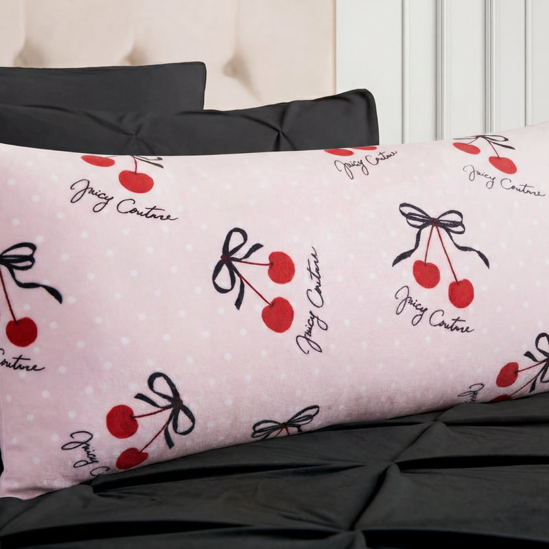 The Plush Body Pillow features a light pink Juicy Couture case with white polka dots, red cherries with black bows, and Juicy Couture script in black, shown on a bed with black pillows and bedspread.