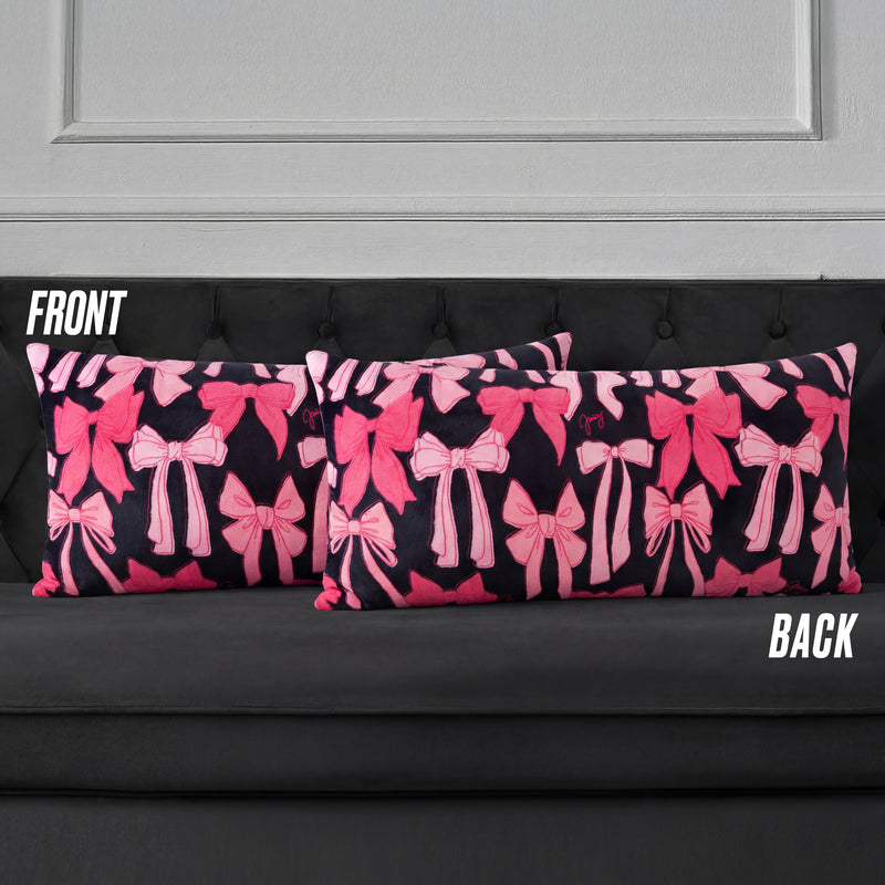 Two rectangular Plush Body Pillows with a pink bow pattern on a dark background are displayed on a black tufted sofa. Labels "FRONT" and "BACK" show both sides feature the same bow design.