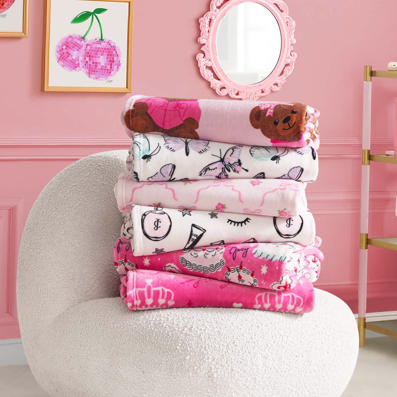 A stack of five ultra-soft blankets, including the Kids Plush Throw Blanket, rests on a plush white chair in a pink room with framed cherry art and a pink mirror on the wall.