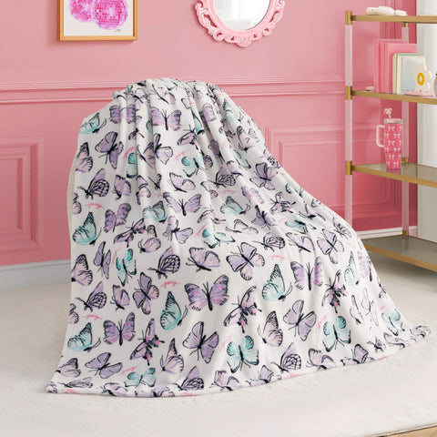 A Kids Plush Throw Blanket with pastel purple, blue, and pink butterfly prints is draped over a chair in a pink room with wall art, a mirror, and gold-accented shelves.