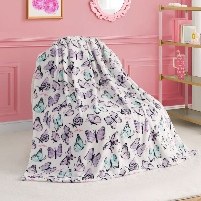 A Kids Plush Throw Blanket with pastel purple, blue, and pink butterfly prints is draped over a chair in a pink room with wall art, a mirror, and gold-accented shelves.