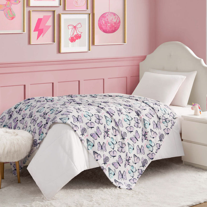 A neatly made bed with a white headboard and butterfly comforter sits in a pink bedroom. Above, framed pictures and a pink disco ball hang. A Kids Plush Throw Blanket adds coziness next to the furry stool and nightstand.