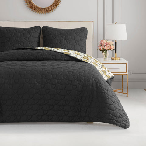 A neatly made bed features a black Cheetah Doodle Hearts Quilt Set with matching pillows and a peek of floral-patterned sheet. The stylish, reversible quilt is machine washable. A white nightstand with a lamp, flowers, and books adds elegance.