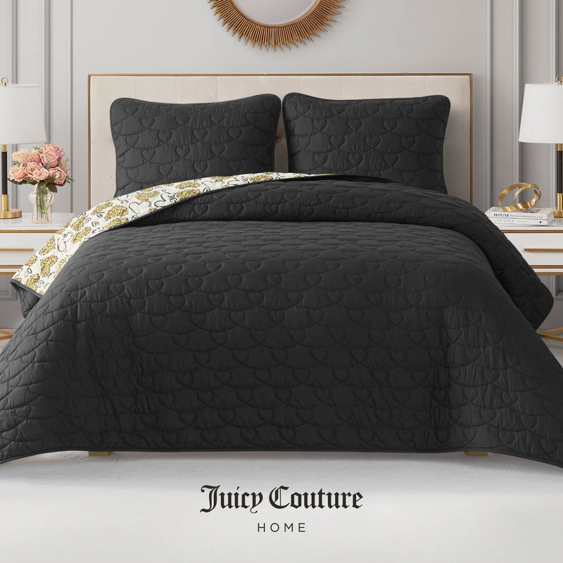 A neatly made bed features the black Cheetah Doodle Hearts Quilt Set with a reversible quilt and matching pillows. Patterned sheets peek out. The room has white walls, nightstands with lamps and flowers, and Juicy Couture Home branding below.
