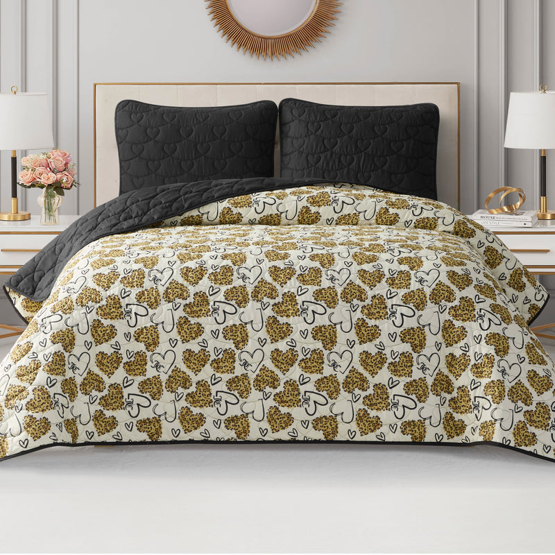 A neatly made bed features the Cheetah Doodle Hearts Quilt Set—a beige and black heart-patterned, reversible, machine-washable quilt—styled with black quilted pillows, modern nightstands, lamps, white walls, and a round decorative mirror.