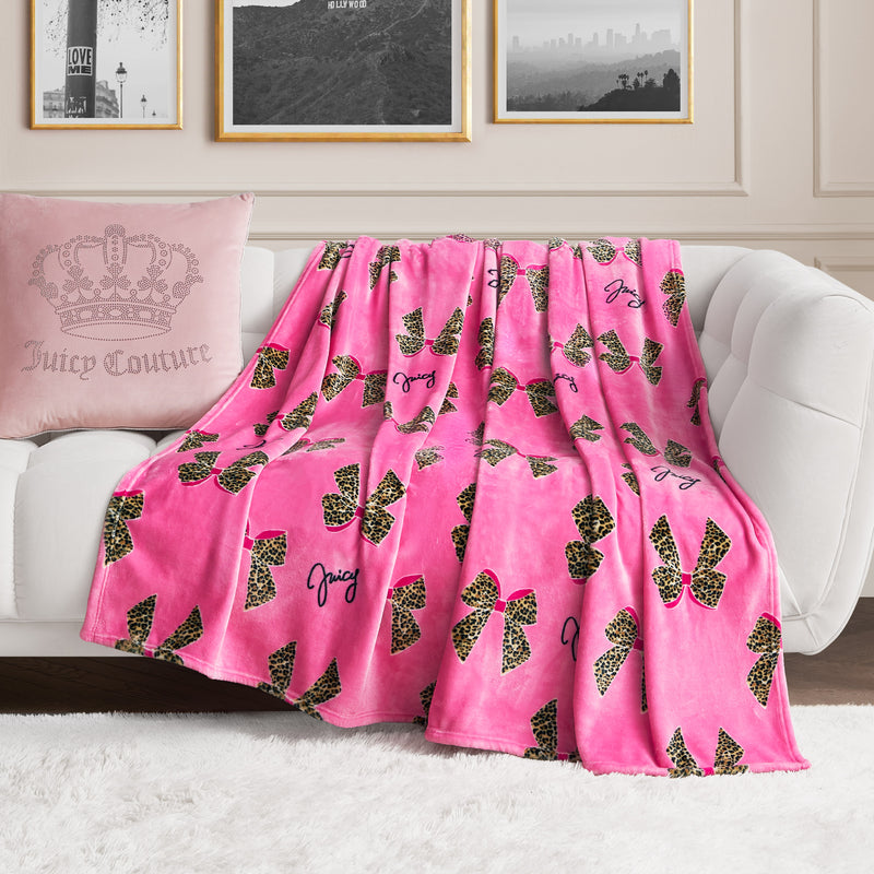 A Plush Oversized Throw Blanket in pink with leopard print bows and Juicy Couture text is draped over a white sofa, complemented by a matching pink pillow with crown and logo accents, adding charm to the elegant living room.