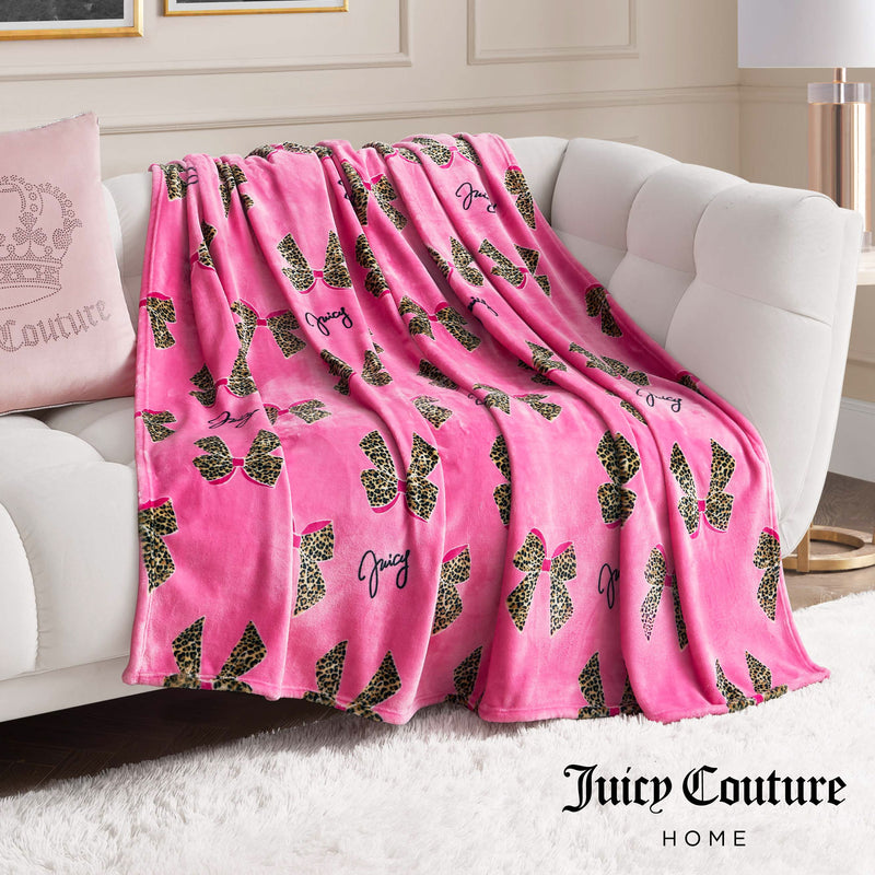The Plush Oversized Throw Blanket in plush pink, decorated with leopard print bows and Juicy text, is draped over a white sofa and paired with a pink crown pillow for stylish flair in a modern living room.
