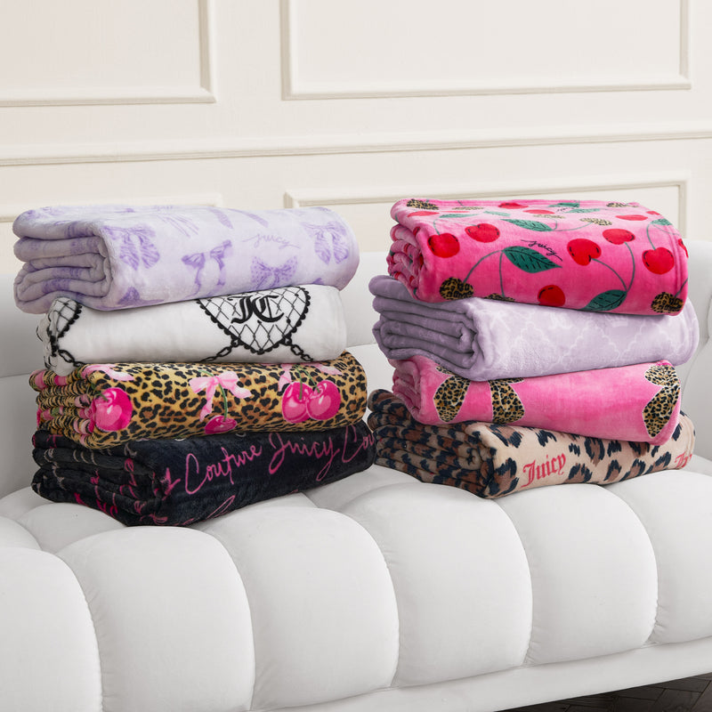 Six folded Plush Oversized Throw Blankets in cherries, leopard print, hearts, and text patterns are neatly stacked on a white tufted bench. Cozy up in style with each ultra-soft blanket against a light-colored wall.