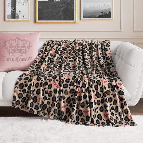 A Plush Oversized Throw Blanket with leopard print and red Juicy Couture text is draped over a white sofa, complemented by a pink Juicy Couture pillow and black-and-white framed photos on the wall behind.