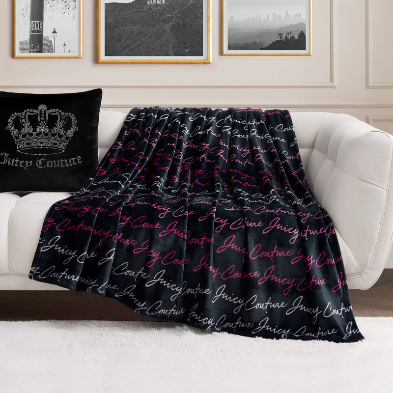 A Plush Oversized Throw Blanket with “Juicy Couture” in white and pink script is draped over a white sofa next to a black Juicy Couture pillow, set in a modern living room with framed wall art.