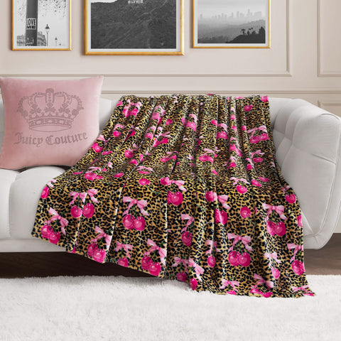 A Plush Oversized Throw Blanket with leopard print and pink cherries is draped over a white sofa with a pink crown pillow, while black and white framed photos decorate the wall.
