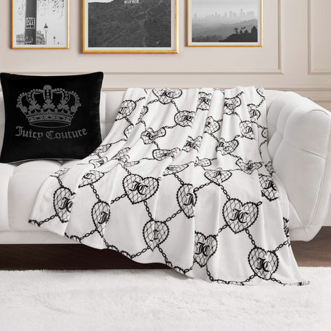 A Plush Oversized Throw Blanket with a black chain and crest pattern is draped over a white couch. Next to it is a black Juicy Couture pillow featuring a crown design. Framed art decorates the beige-paneled wall behind the couch.