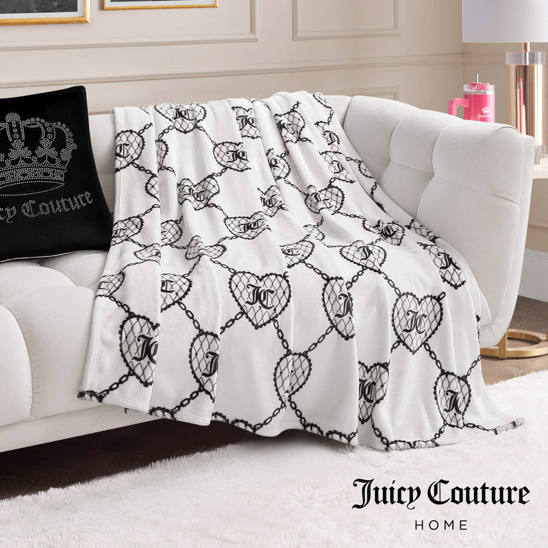 A white couch features a Plush Oversized Throw Blanket by Juicy Couture Home, with linked heart motifs and the letter J. Nearby are a black crown pillow and a pink tumbler, all highlighting the Juicy Couture Home brand.