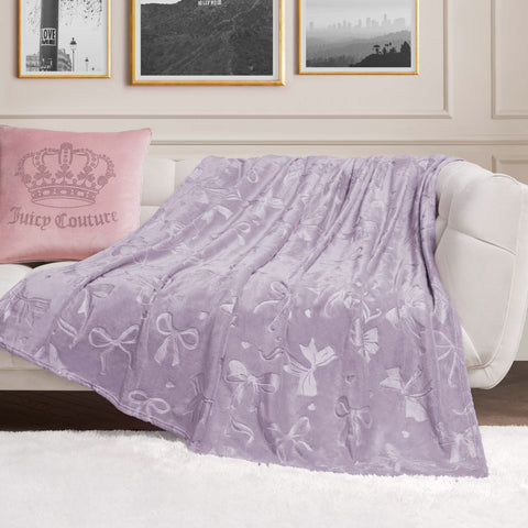 An Embossed Plush Oversized Throw Blanket with soft lavender bow patterns is draped over a white sofa beside a pink Juicy Couture pillow, with black-and-white photos framed on the cream-colored wall behind.