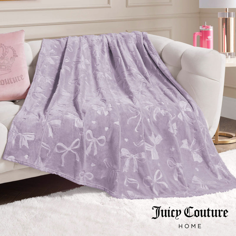 An Embossed Plush Oversized Throw Blanket in light purple with a bow pattern is draped on a white sofa. Nearby are a pink Juicy Couture pillow and tumbler. The Juicy Couture Home logo appears at the bottom right.