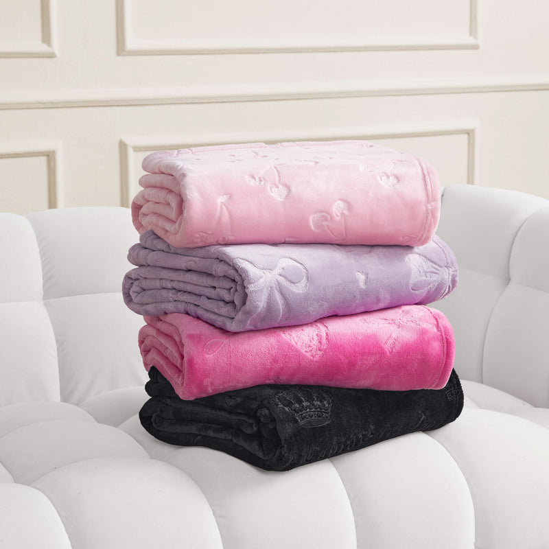 Four folded Embossed Plush Oversized Throw Blankets in pink, lavender, hot pink, and black are stacked on a white tufted sofa. Each features a subtle embossed bow pattern. The background shows a cream wall with decorative molding.