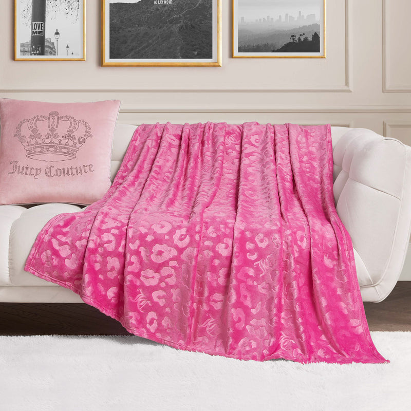 An Embossed Plush Oversized Throw Blanket in pink leopard print is draped over a white sofa, paired with a light pink "Juicy Couture" pillow. Framed photos and crisp white paneled walls complete the chic scene.