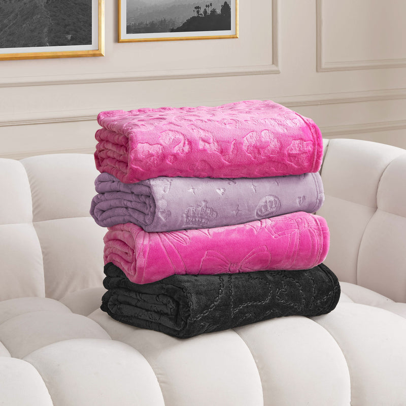 Four folded Embossed Plush Oversized Throw Blankets in pink, lavender, fuchsia, and black are stacked on a white tufted sofa. Framed wall art adds a finishing touch to the cozy scene.
