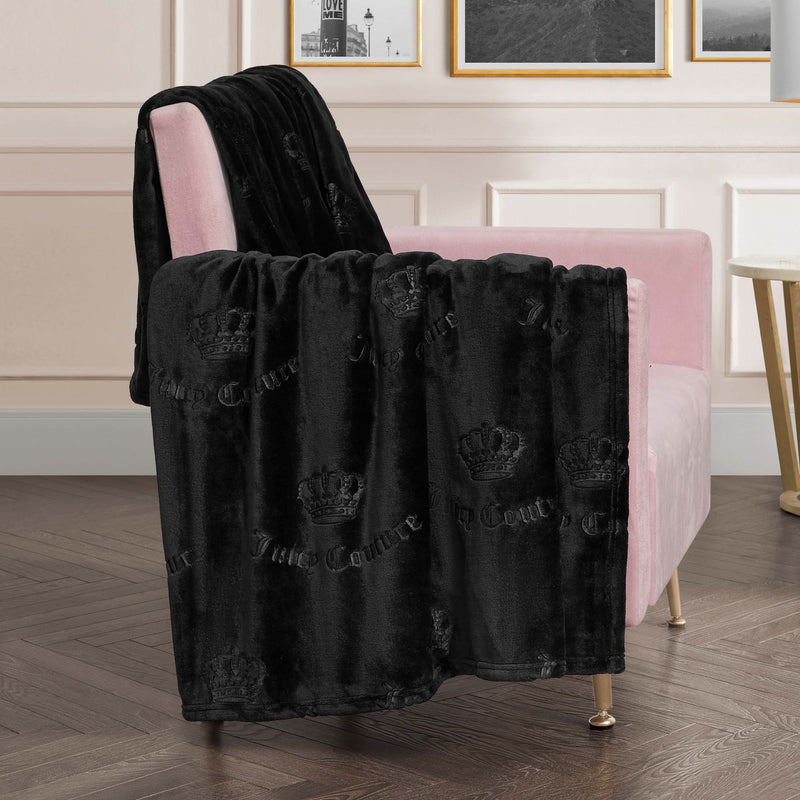 An Embossed Plush Oversized Throw Blanket from Juicy Couture with a crown pattern is draped over a light pink armchair in a chic modern room with white paneled walls, framed art, and a wooden herringbone floor.