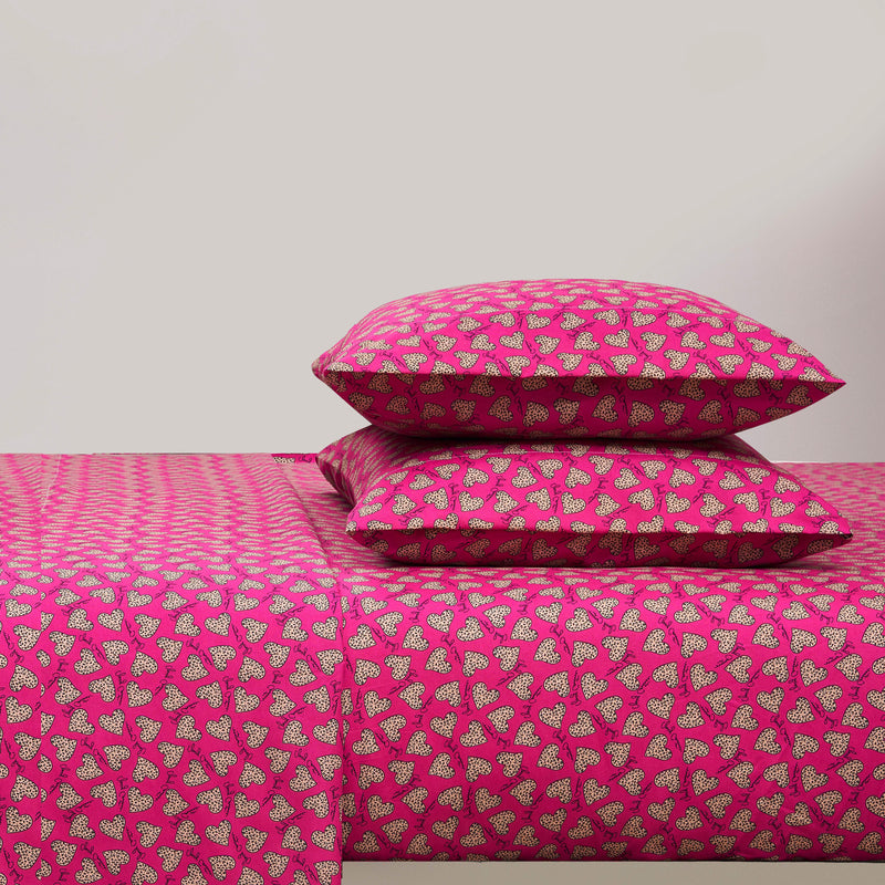 A bed with a bright pink Printed Microfiber Sheet Set and two matching pillows, all featuring a repeating pattern of small white and black hearts. The background wall is plain and light-colored.