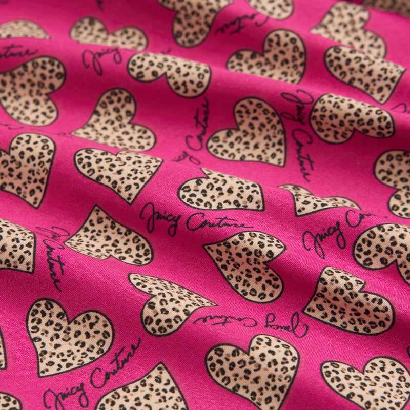 A close-up of pink fabric from the Printed Microfiber Sheet Set shows leopard-print hearts and "Juicy Couture" in black script. The slightly wrinkled microfiber gives this luxury bedding a relaxed, stylish appearance.