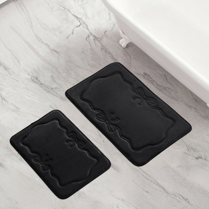 Two black Quilted Memory Foam Bath Mats featuring decorative edges and bow designs are placed on a light gray marble bathroom floor beside a white bathtub.