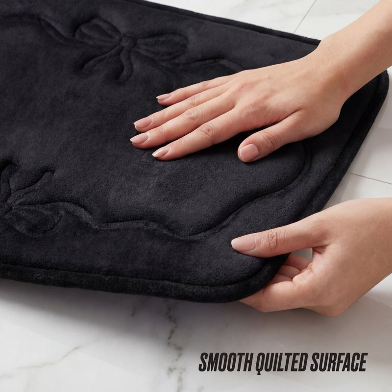 Two hands touch a black Quilted Memory Foam Bath Mat 2-Piece Set with a raised bow design on a white marble surface. The mat’s soft quilted texture stands out. “SMOOTH QUILTED SURFACE” text appears in the corner.