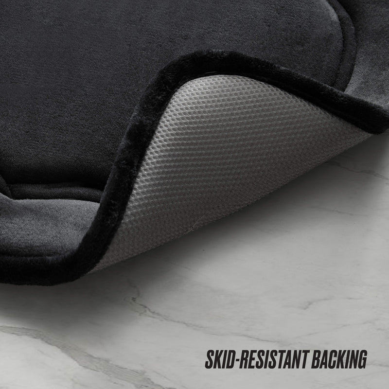 A close-up of the Quilted Memory Foam Bath Mat 2-Piece Set in dark gray, with a corner lifted to show its textured, skid-resistant light gray backing on a light marble-like floor. Text: SKID-RESISTANT BACKING.