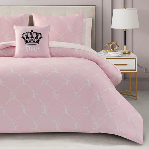 A neatly made bed with the Link Printed Bed in a Bag Bedding Set in light pink, featuring a white JC pattern, paired with a matching Juicy pillow with black crown and text, next to a modern white nightstand holding a lamp, book, and gold glasses.