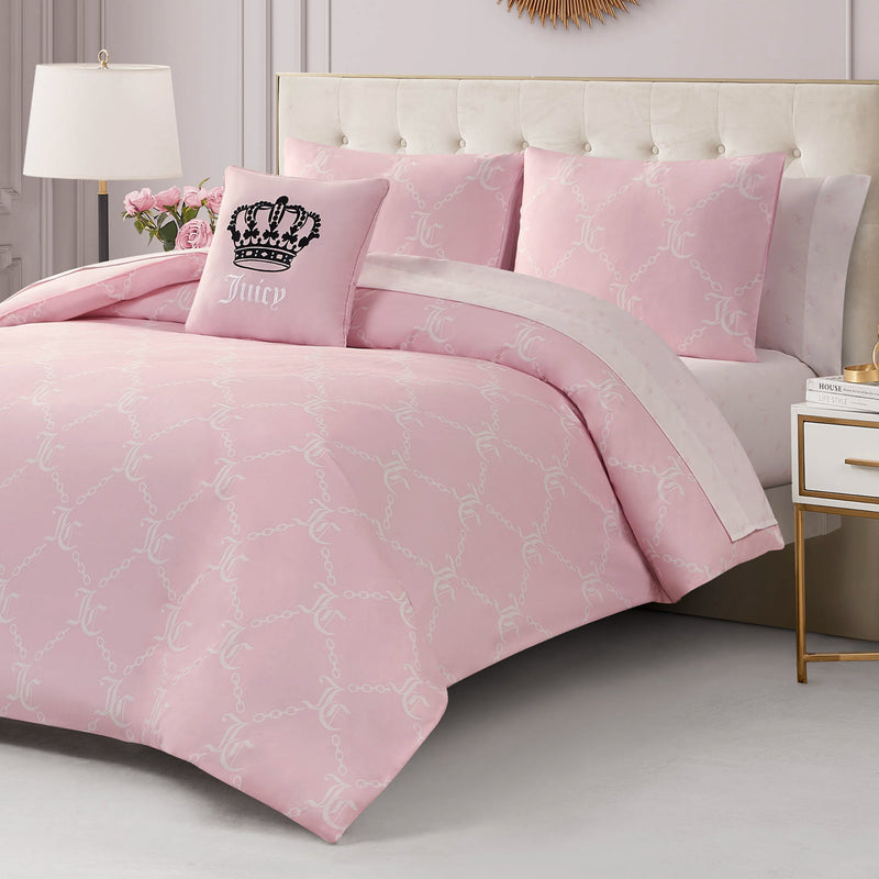 A neatly made bed features the Link Printed Bed in a Bag Bedding Set in light pink with a subtle pattern, matching pillows, and a decorative crown pillow reading Juicy. The room is styled with a white nightstand, lamp, and pink flowers for added luxury.
