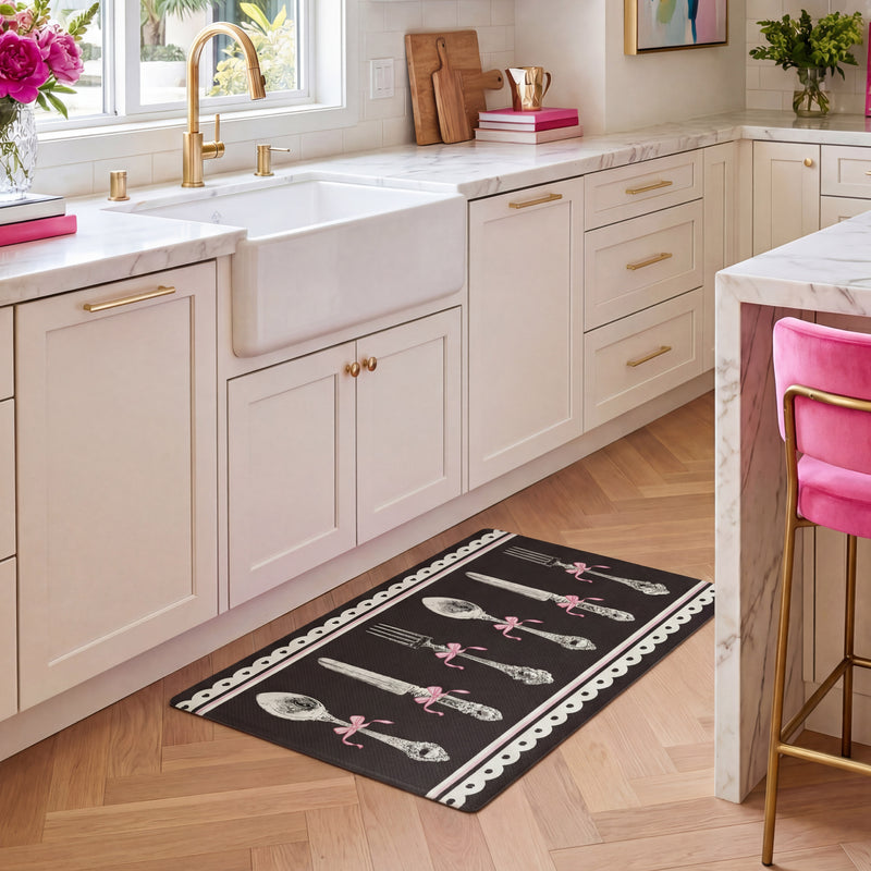A modern kitchen features white cabinets, gold hardware, and a white farmhouse sink. The Kitchen Wellness Mat with vintage silverware designs lies on the wooden floor, offering comfort and support as an anti-fatigue mat. Pops of color come from pink flowers and a pink chair in the space.