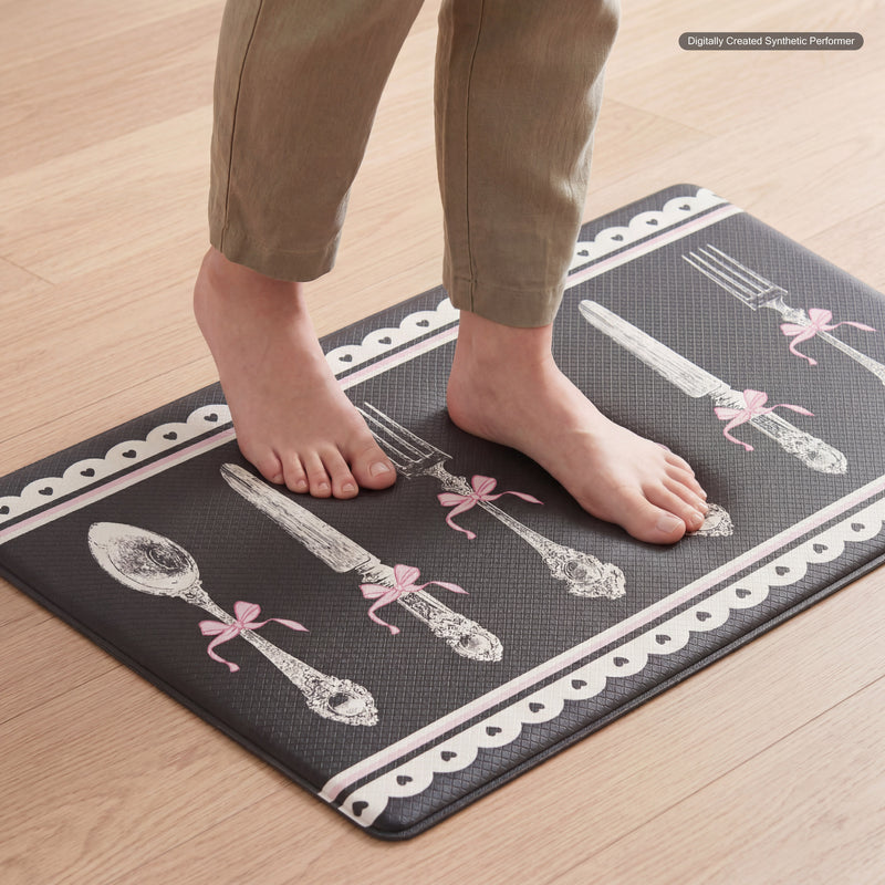 A person stands barefoot on a Kitchen Wellness Mat, a stylish floor mat adorned with vintage silverware illustrations and pink bows. This anti-fatigue mat rests on a light wooden floor, blending comfort with chic design.