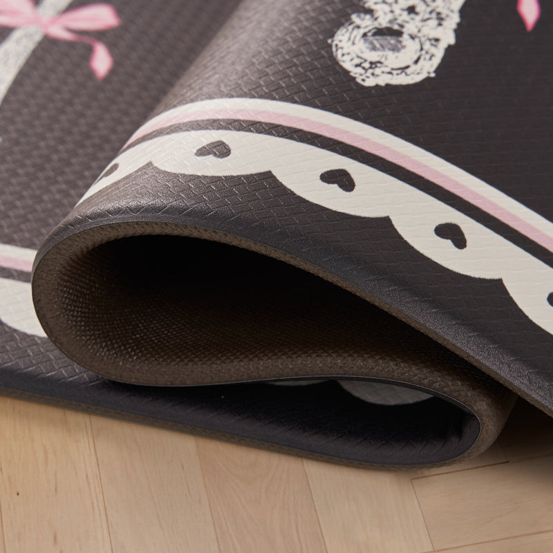 A close-up of a rolled Kitchen Wellness Mat on a wooden floor, featuring a scalloped white border with small black hearts and pink accents on a dark background. This stylish anti-fatigue mat from Juicy Couture adds comfort and charm to any space.
