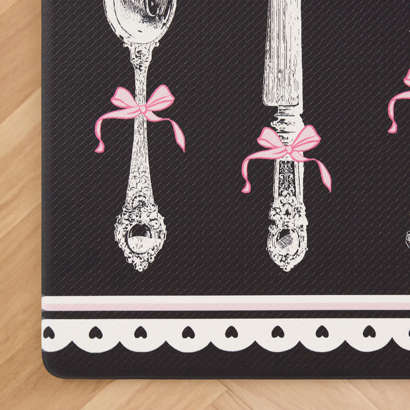 Illustration of ornate silverware adorned with pink bows set against a black background, accented by a white scalloped border with black dots along the bottom edge for added elegance. The design is placed on a wood floor, making it ideal as the Kitchen Wellness Mat—a stylish and anti-fatigue floor mat inspired by Juicy Couture Wellness Mat aesthetics.