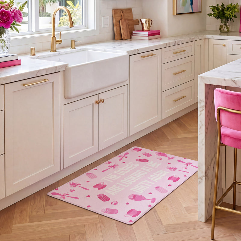 A modern kitchen with white cabinets, gold hardware, and a farmhouse sink. The Kitchen Wellness Mat, featuring playful cupcake prints and uplifting text, sits on the floor as a stylish and comfortable anti-fatigue mat. Pink barstool and flowers add cheerful pops of color to the space.