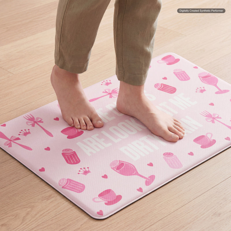 Bare feet stand on a pink Kitchen Wellness Mat from Juicy Couture, which features white and pink kitchen-themed illustrations and hearts. This anti-fatigue mat displays a partially visible message in white text and is placed on a light wood kitchen floor.