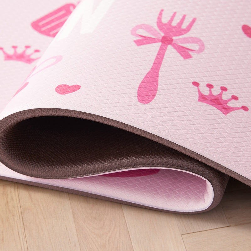 A close-up of a rolled Kitchen Wellness Mat featuring playful prints of crowns, hearts, a bow, and a fork, placed on a light wooden floor.