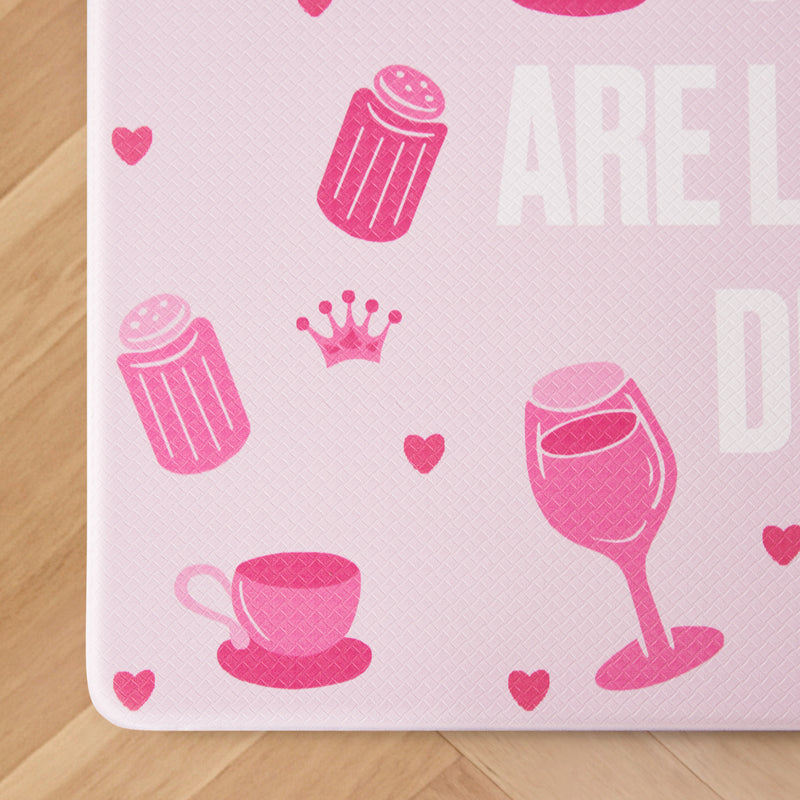 Pink icons of a teacup, wine glass, salt shaker, crown, and small hearts are scattered across a textured light pink surface with partial large white text on the right side, bringing a playful vibe to this Kitchen Wellness Mat—a stylish anti-fatigue mat designed to brighten up any kitchen floor.