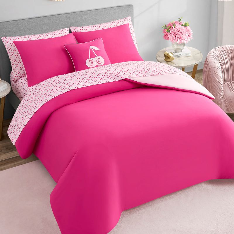 A neatly made bed features the Cherry Reversible Bed in a Bag Bedding Set, matching pillows, and a decorative cherry-embroidered pillow. Facing a gray headboard, the bed is next to a side table with pink flowers in a vase.