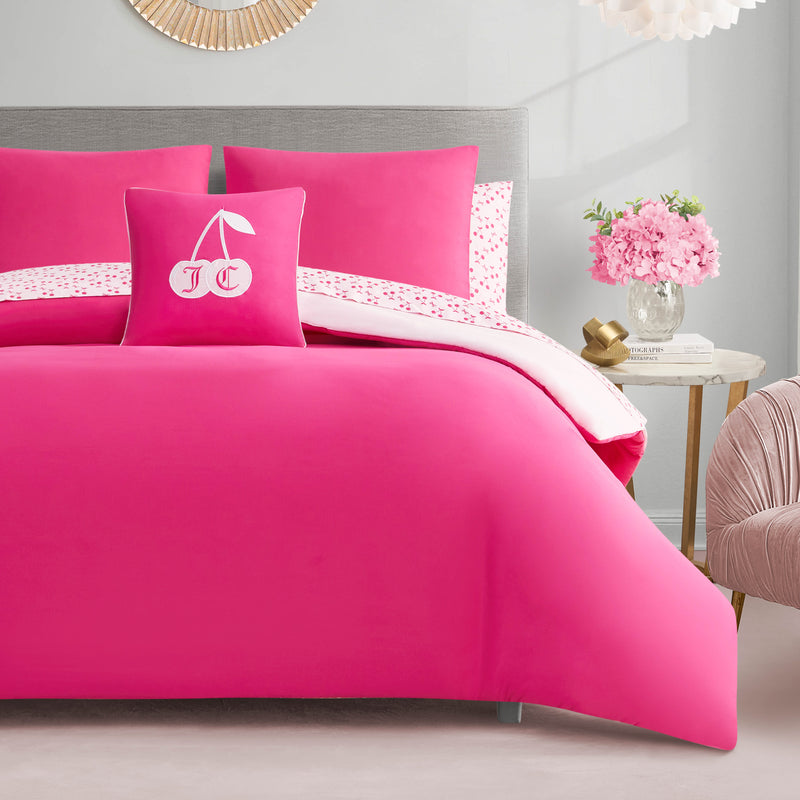 A neatly made bed with the Cherry Reversible Bed in a Bag Bedding Set brings style to bright pink bedding and matching pillows. The center pillow has embroidered cherries and JC initials. A nightstand with pink flowers sits by neutral walls.