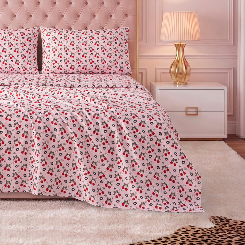 A neatly made bed with a light pink tufted headboard, red and black floral luxury bedding, matching pillows, and a Printed Microfiber Sheet Set. Beside the bed are a gold lamp and white nightstand on a plush white rug.